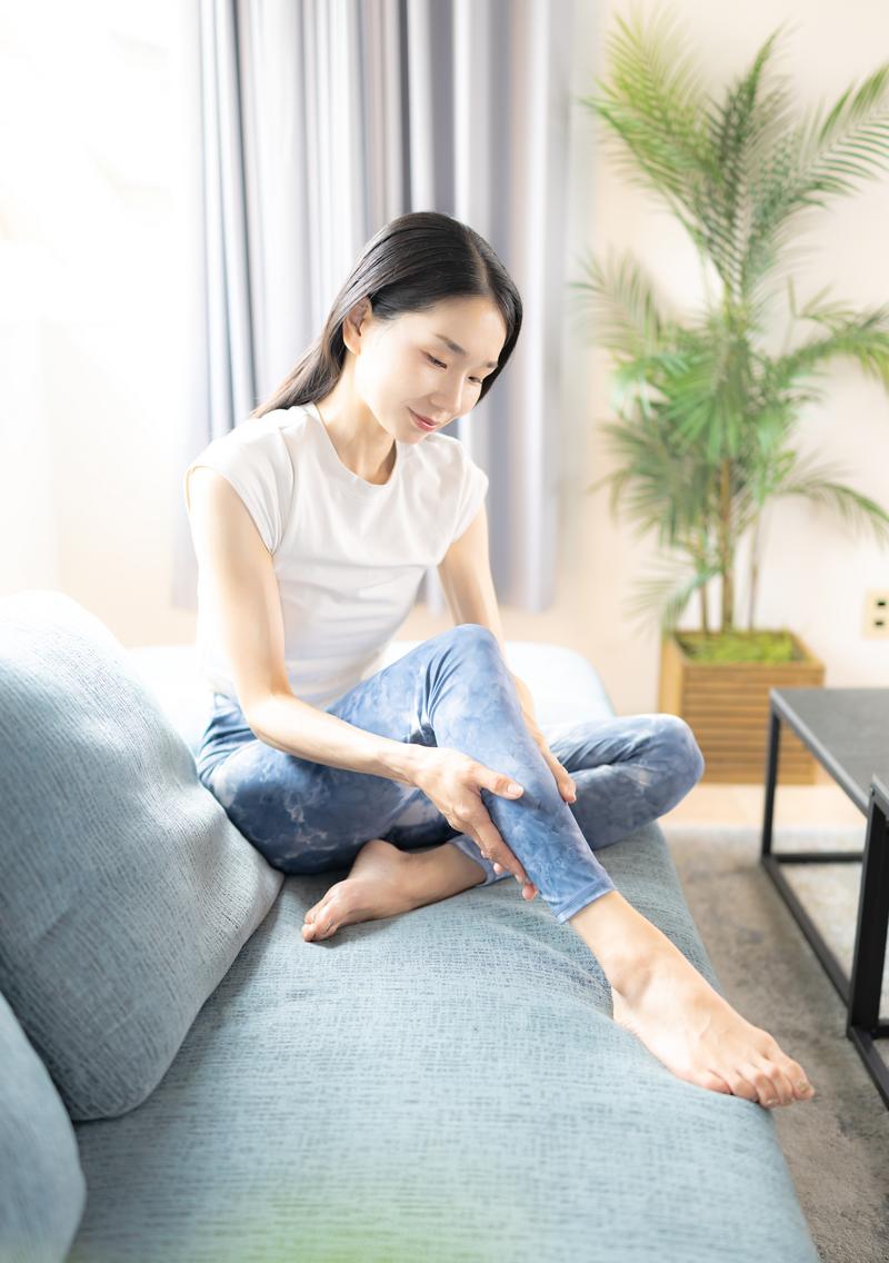 自宅のソファで、脚に手を添えてストレッチをしている女性(モデル)