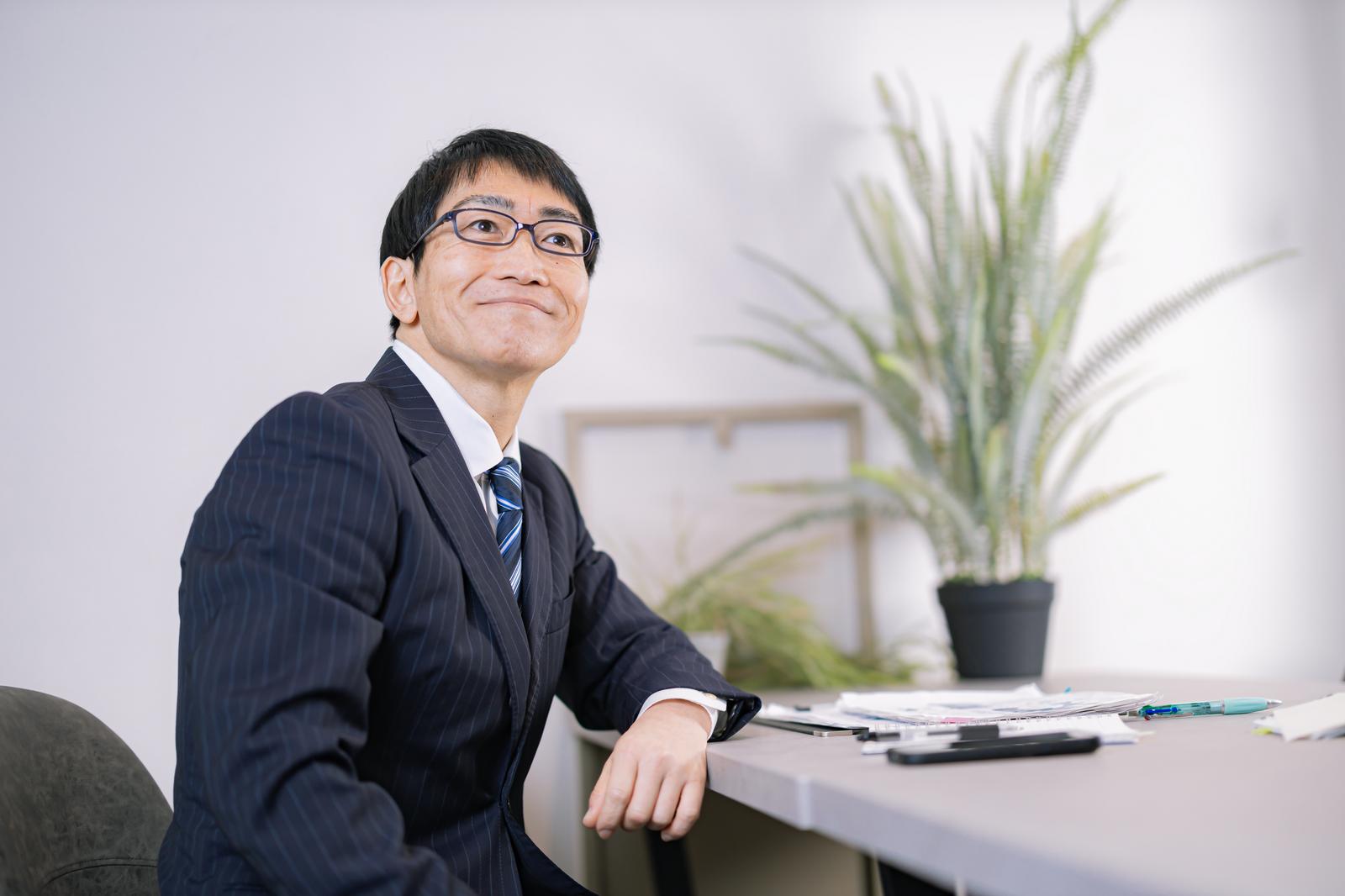 代表のイメージ写真(モデル)。事務所のデスクに座り、ほほえむスーツ姿の男性
