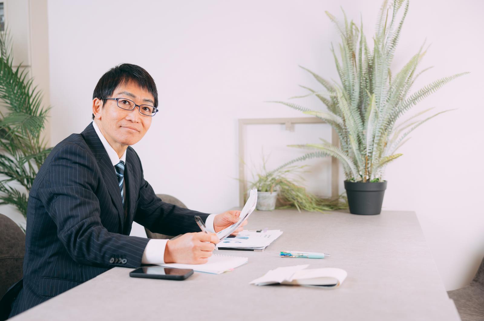 代表のイメージ写真(モデル)。事務所のデスクで書類を手に、こちらを向いてほほえむスーツ姿の男性