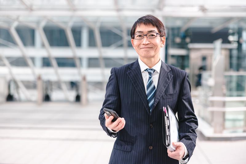 代表のイメージ写真(モデル)。バインダーを抱え、屋外でほほえむスーツ姿の男性